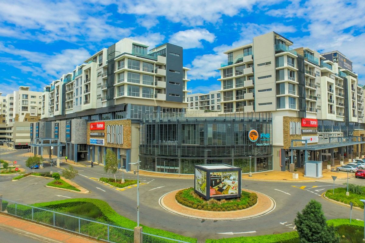 Aerial view of a modern multi-story apartment complex featuring ground-level commercial retail spaces, paved access roads, and landscaped surroundings.