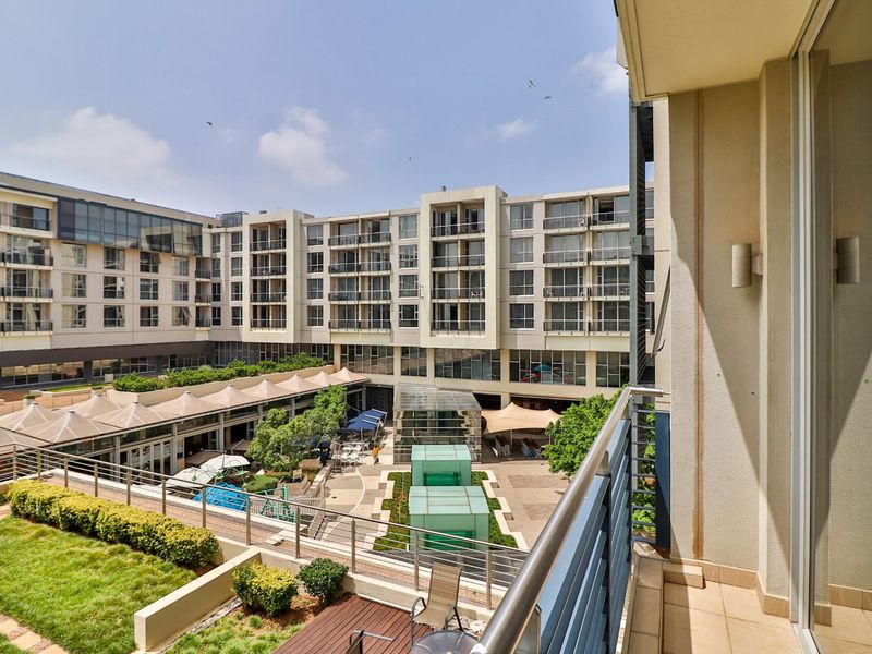 A private balcony featuring tiled flooring and a stainless steel balustrade, offering an elevated view over the apartment complex courtyard and landscaped gardens.