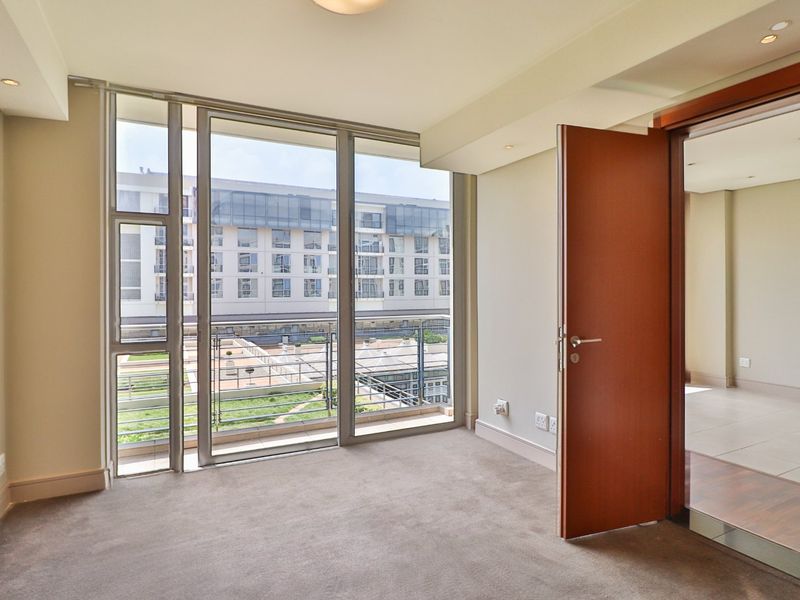 A bright, empty bedroom featuring floor-to-ceiling windows with balcony access, neutral carpeting, and recessed lighting, offering a clean and modern living space.