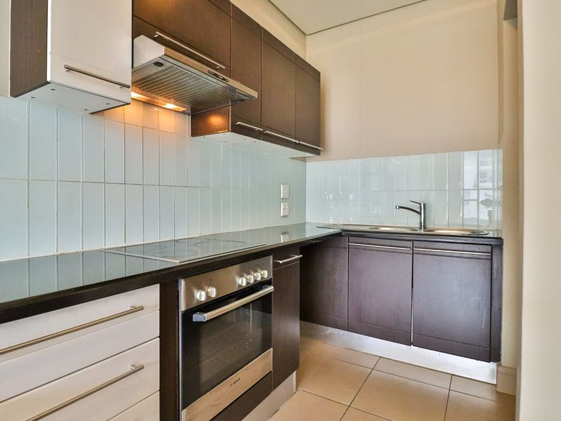 A modern, compact kitchen featuring dark wood cabinetry, a tiled backsplash, built-in oven, electric cooktop, and stainless steel sink.