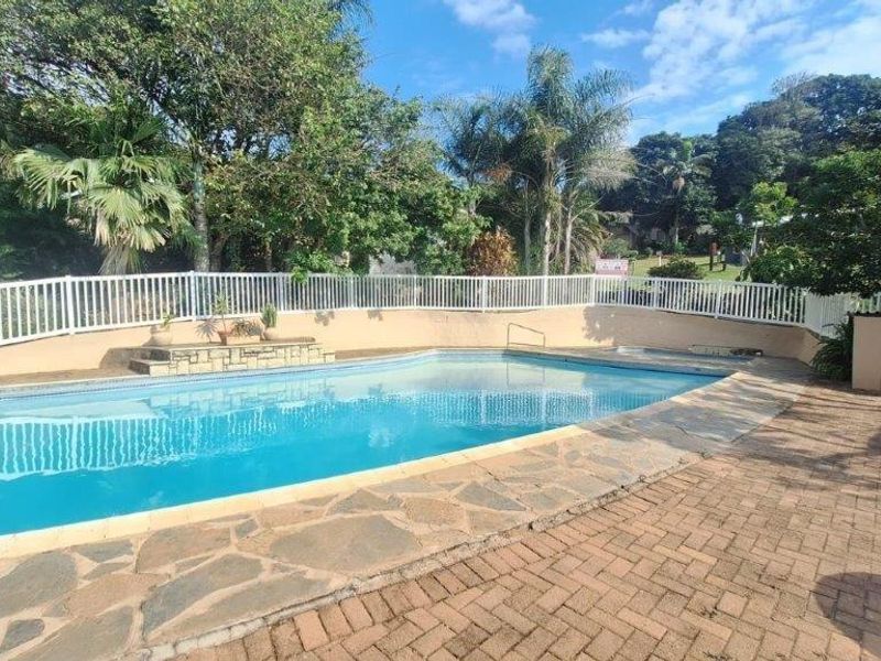 A well-maintained outdoor swimming pool area featuring stone and brick paving, perimeter fencing, and established garden surroundings.