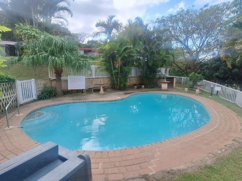 A communal outdoor swimming pool area featuring brick coping, perimeter fencing, and established garden surroundings within the townhouse complex.