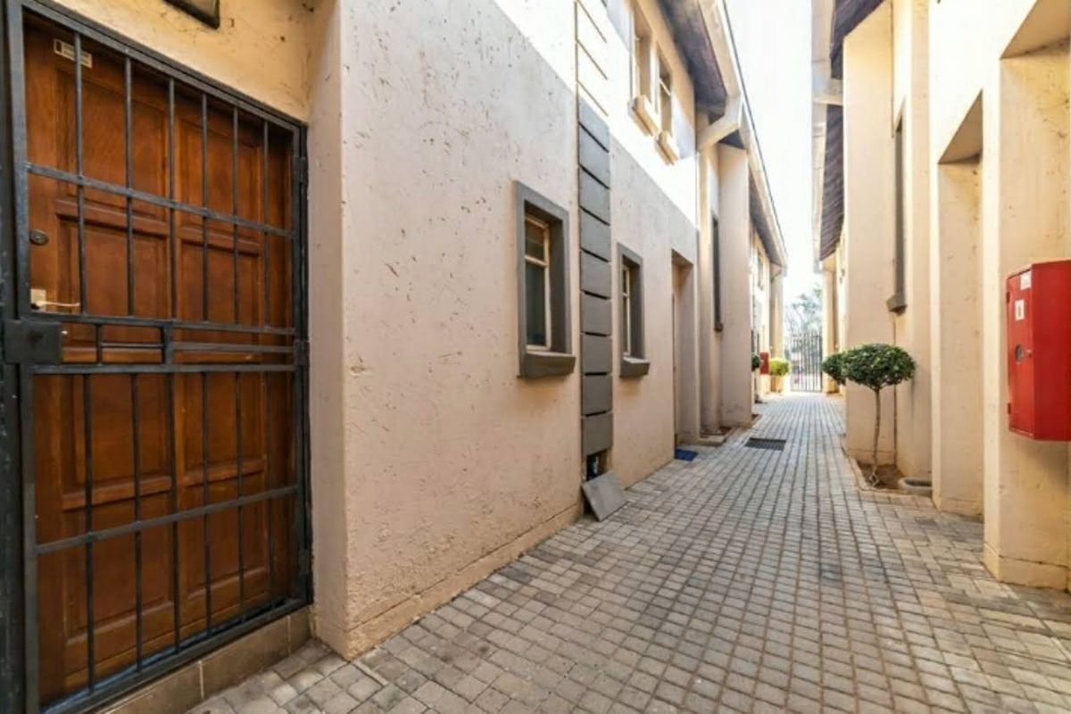 A narrow exterior paved walkway between apartment building sections, featuring a security gate and wall-mounted fire safety equipment.