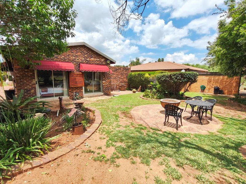 A well-maintained rear garden featuring a face brick facade, paved circular patio area, and established landscaping with retractable awnings over sliding glass doors.