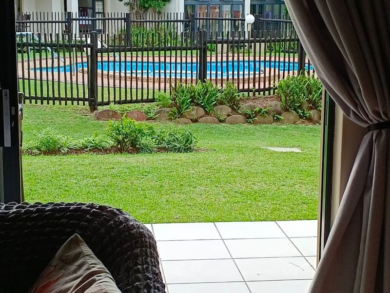 A view from a tiled patio looking out onto a communal garden area with a swimming pool and perimeter fencing.