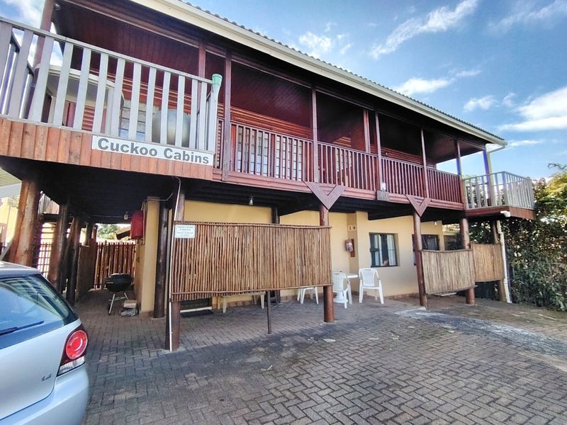 Front elevation of a two-story timber-clad cabin featuring a raised balcony, paved driveway, and stilt-supported structure.