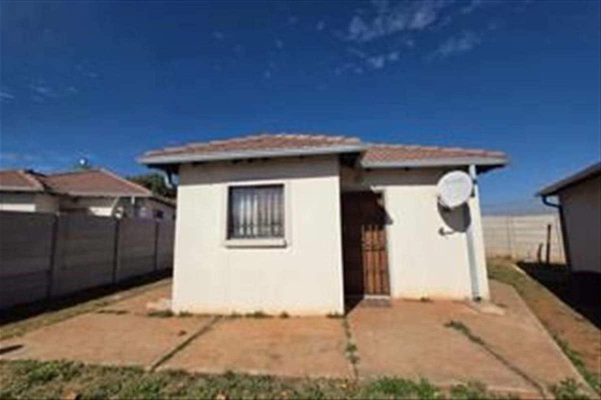 Rear view of a detached house featuring a concrete patio, tiled roof, and secure window bars, set within a fenced yard.