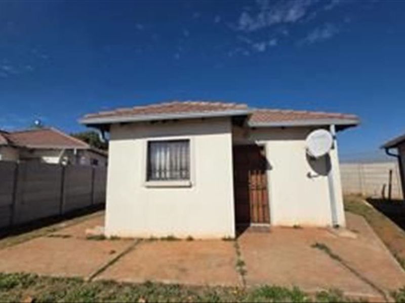 Rear view of a detached house featuring a concrete patio, tiled roof, and secure window bars, set within a fenced yard.