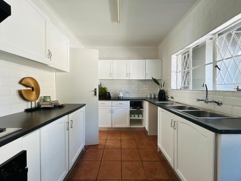 A functional kitchen featuring white cabinetry, dark countertops, subway tile backsplash, and terracotta floor tiles with a double sink and window security bars.
