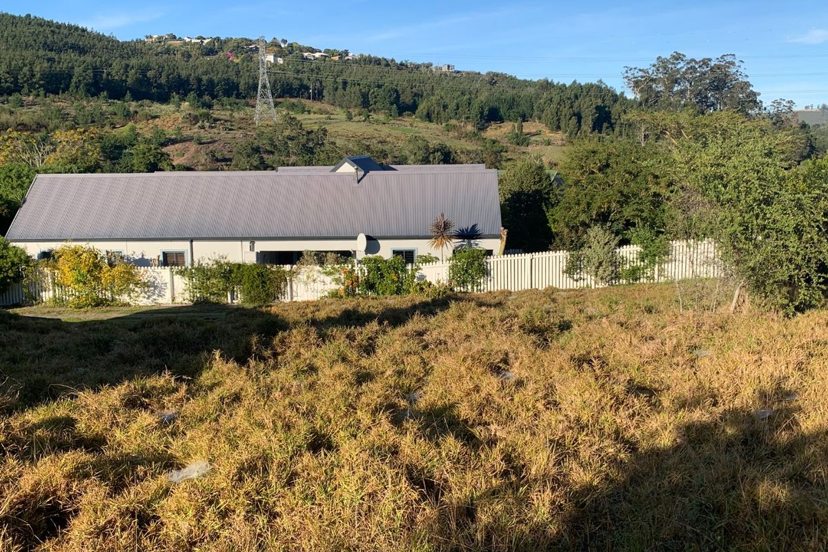 A view of a vacant residential plot with natural grass, bordered by a white fence and overlooking a neighboring property with a metal roof.