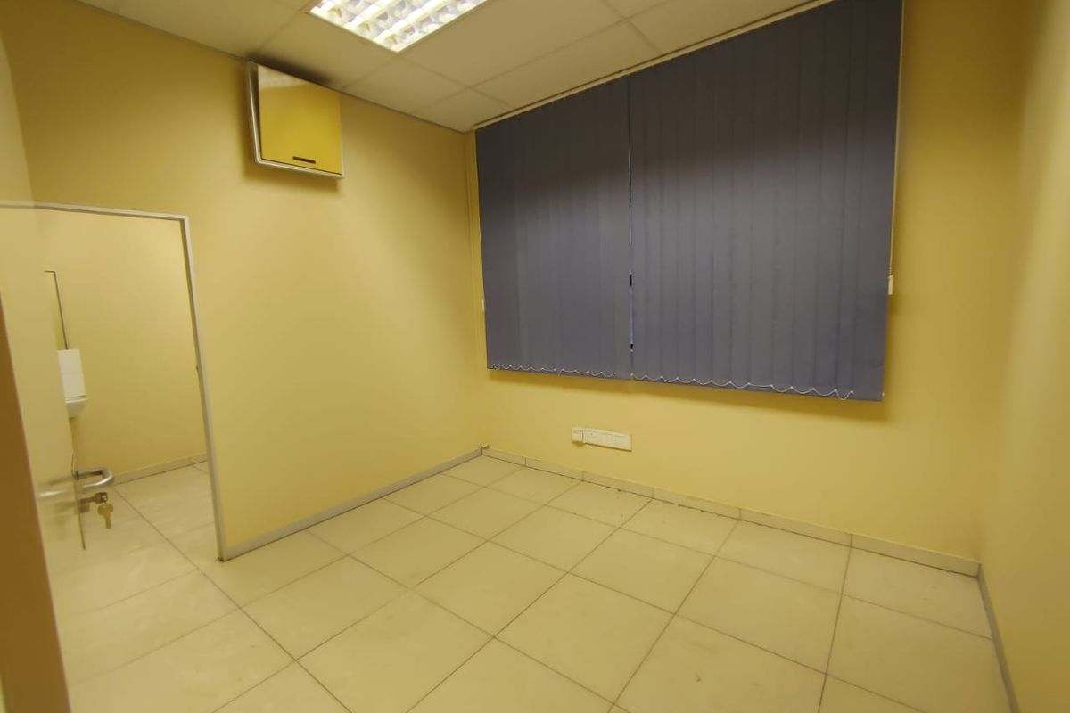 An empty office space featuring tiled flooring, a suspended ceiling with recessed lighting, and vertical window blinds.