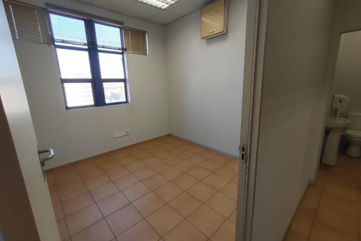 An empty commercial office space featuring tiled flooring, a suspended ceiling with recessed lighting, and a wall-mounted air conditioning unit.