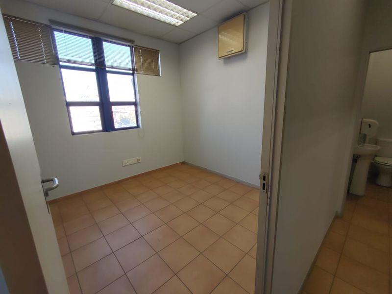 An empty commercial office space featuring tiled flooring, a suspended ceiling with recessed lighting, and a wall-mounted air conditioning unit.