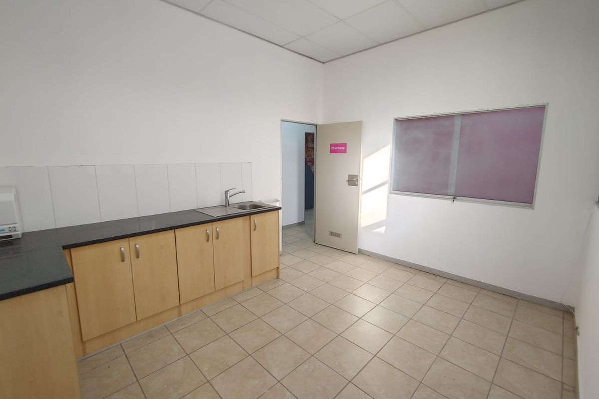 A functional commercial kitchenette featuring light wood cabinetry, dark laminate countertops, a stainless steel sink, and neutral tiled flooring.