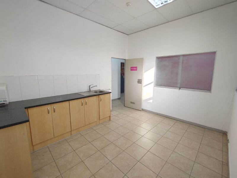 A functional commercial kitchenette featuring light wood cabinetry, dark laminate countertops, a stainless steel sink, and neutral tiled flooring.