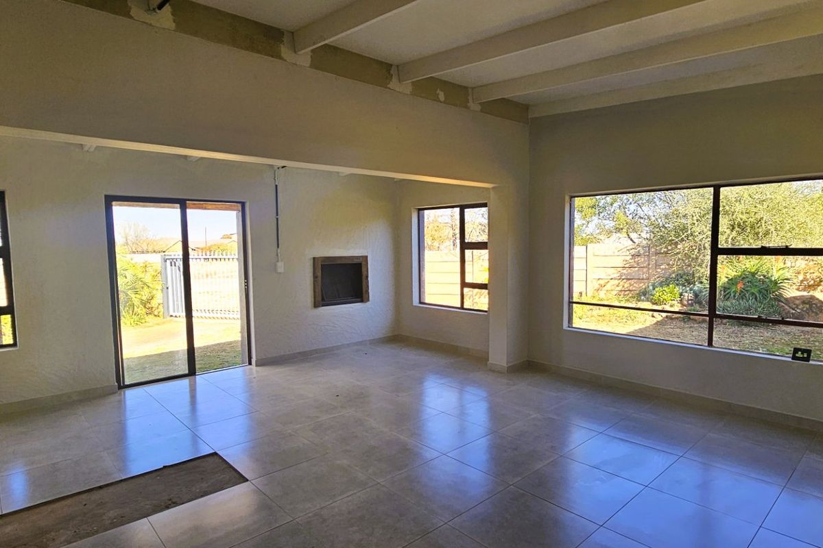 Spacious, empty living area featuring tiled floors, exposed ceiling beams, a built-in fireplace, and large windows with garden views.
