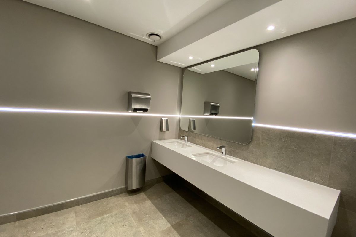Modern commercial bathroom featuring a sleek double vanity with integrated sinks, large mirror, and ambient LED strip lighting.