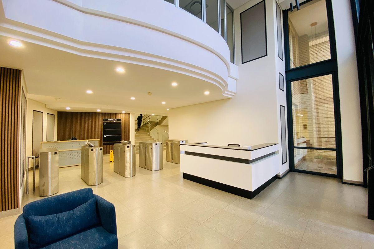 Modern commercial building lobby featuring a reception desk, security turnstiles, high ceilings, and large glass windows providing ample natural light.