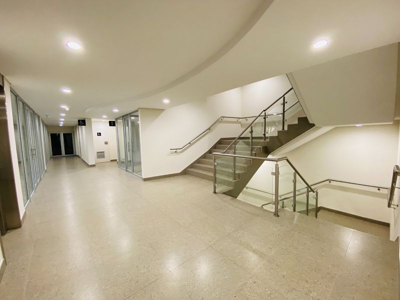 Modern commercial hallway featuring tiled flooring, recessed lighting, and a staircase with a contemporary glass balustrade and stainless steel handrails.