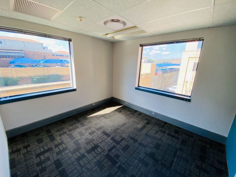 A vacant, well-lit office space featuring carpeted flooring, a suspended ceiling with recessed lighting, and large windows providing natural light.