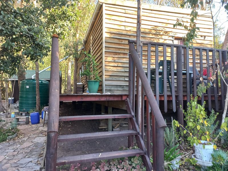 A rustic timber cabin featuring a raised deck, wooden staircase, and an adjacent rainwater tank set within a natural, wooded environment.