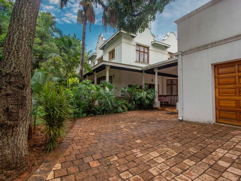 A charming multi-story home featuring a brick-paved driveway, a wooden garage door, and a covered patio area surrounded by established garden landscaping.