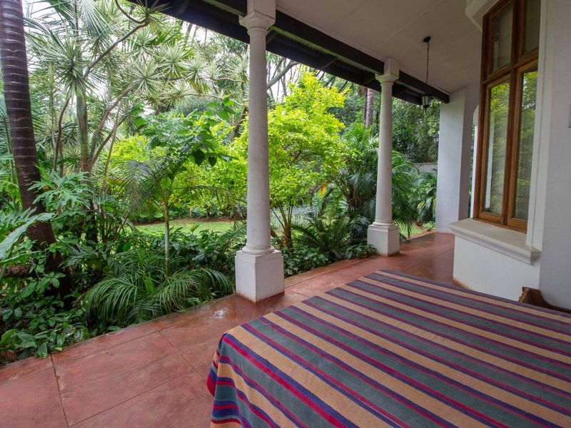 A covered patio featuring classical columns and tiled flooring, overlooking a lush, established garden setting.