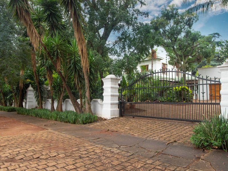 Front entrance view featuring a secure wrought iron gate, masonry pillars, and a paved driveway surrounded by mature landscaping.