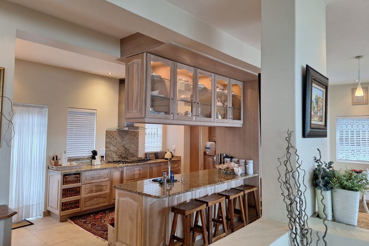 A well-appointed kitchen featuring wooden cabinetry, granite countertops, a central island with sink, and elegant hanging glass-front display cabinets with integrated lighting.