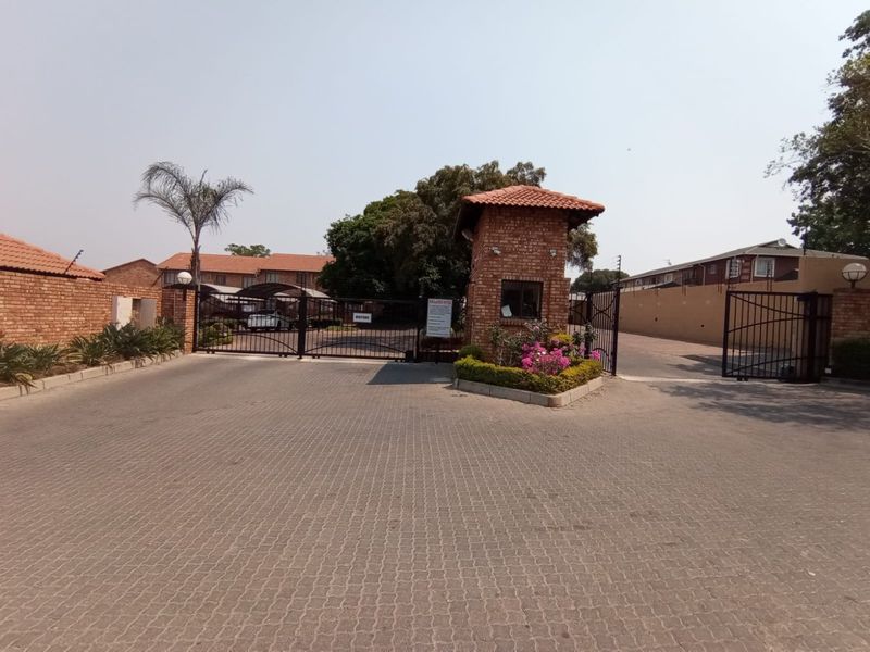The entrance to the townhouse complex features a brick security guard house, wrought iron gates, and a paved driveway with perimeter security fencing.