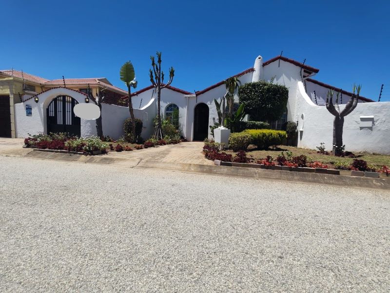 Front elevation of a white stucco house featuring an arched gate, paved driveway, and established garden landscaping under a clear blue sky.