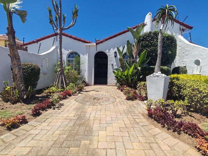 A well-maintained front entrance featuring a paved walkway, arched doorway with security gate, and established garden landscaping under a tiled roof.
