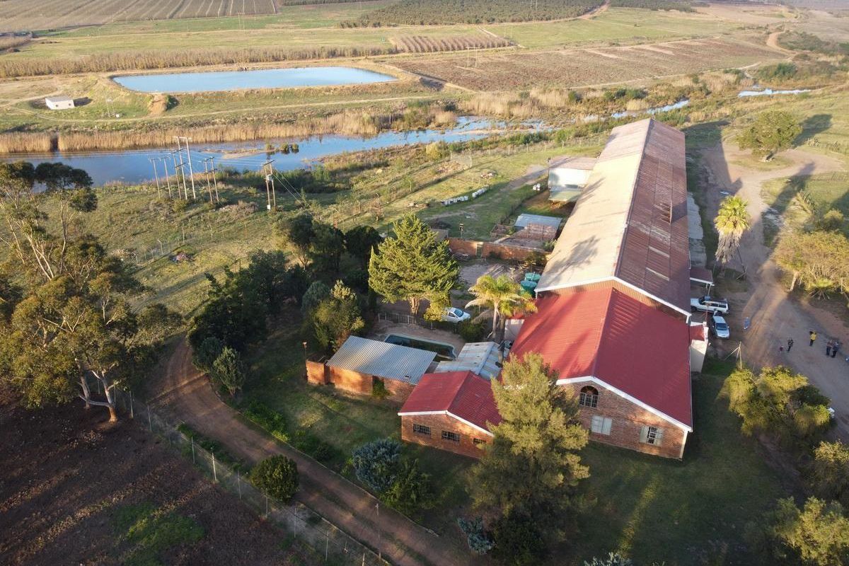 Aerial view of a farm property featuring a large brick building with a metal roof, swimming pool, and surrounding agricultural land with a dam.