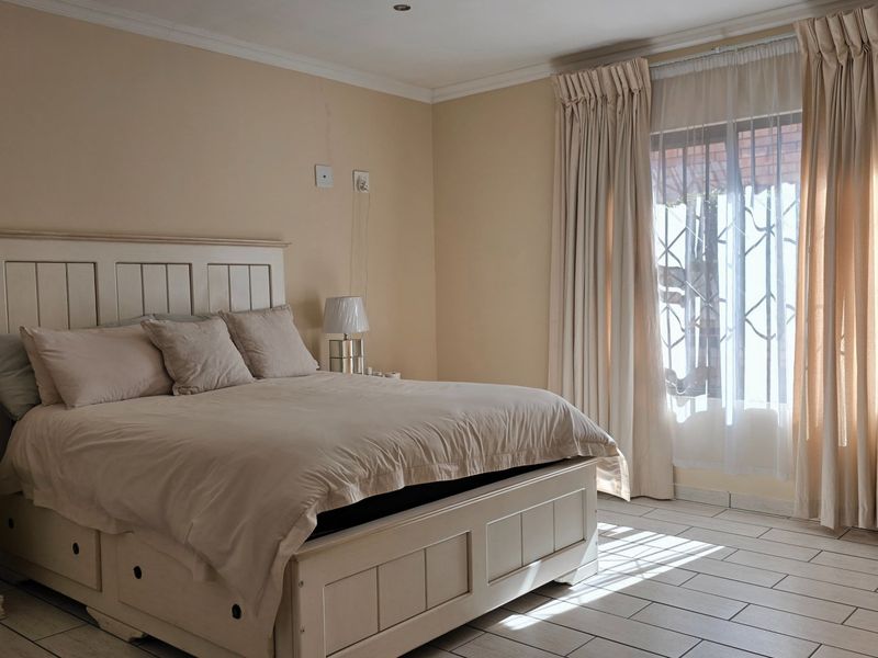 A well-lit bedroom featuring neutral wall tones, tiled flooring, and large windows fitted with security gates and curtains.