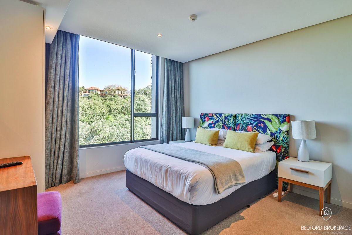 A well-lit bedroom featuring large floor-to-ceiling windows with natural views, neutral carpeted flooring, and recessed ceiling lighting.