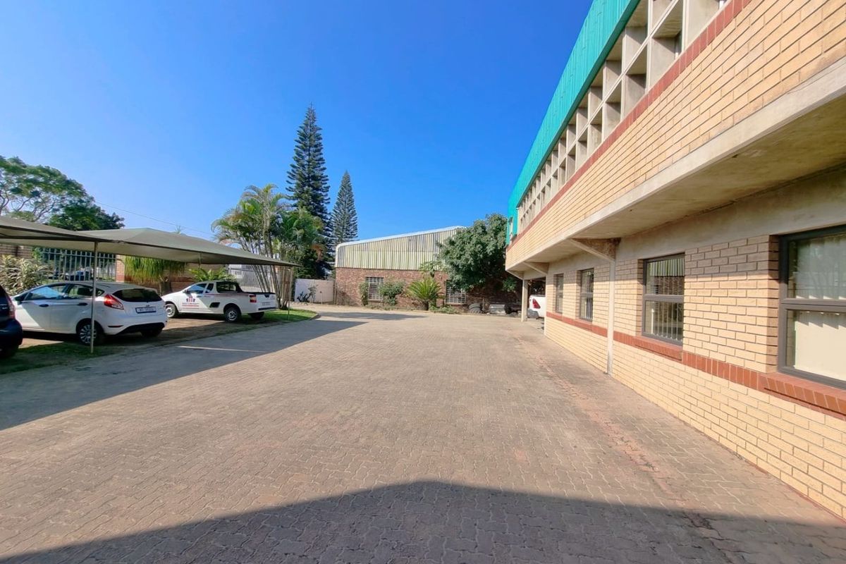 Exterior view of a commercial property featuring a paved driveway, covered parking area, and brick building facade with security window bars.