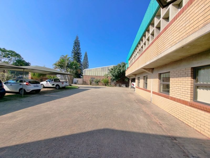 Exterior view of a commercial property featuring a paved driveway, covered parking area, and brick building facade with security window bars.