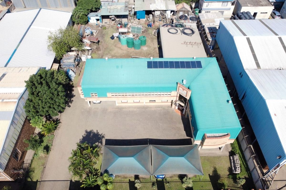 Aerial view of a commercial property featuring an L-shaped building with a metal roof, solar panels, paved parking area, and shade cloth carports.