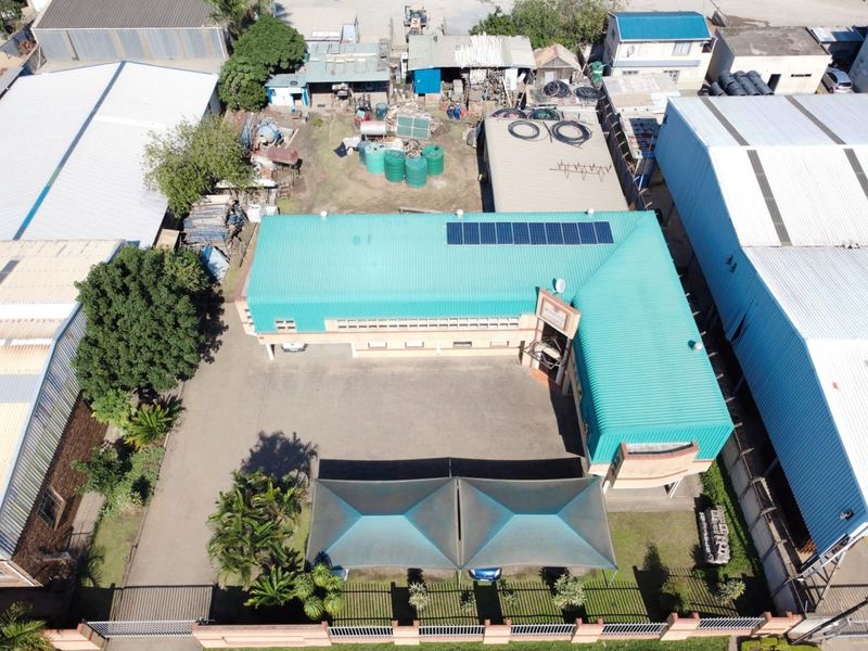 Aerial view of a commercial property featuring an L-shaped building with a metal roof, solar panels, paved parking area, and shade cloth carports.