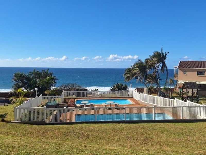A communal swimming pool area featuring a paved deck, secure perimeter fencing, and expansive lawn, set against a scenic ocean view.