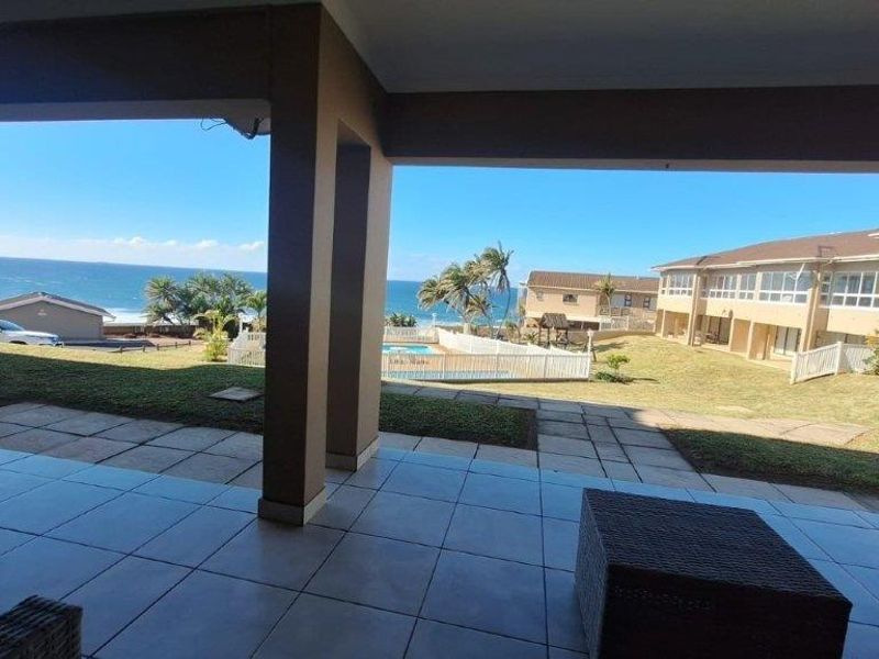 A covered patio area featuring tiled flooring and support columns, offering an open view of the communal garden, swimming pool, and ocean.
