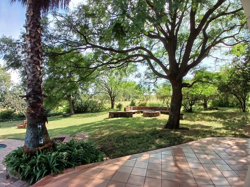 A spacious garden area featuring a stone fire pit with built-in bench seating, mature trees, and a tiled patio overlooking the landscape.