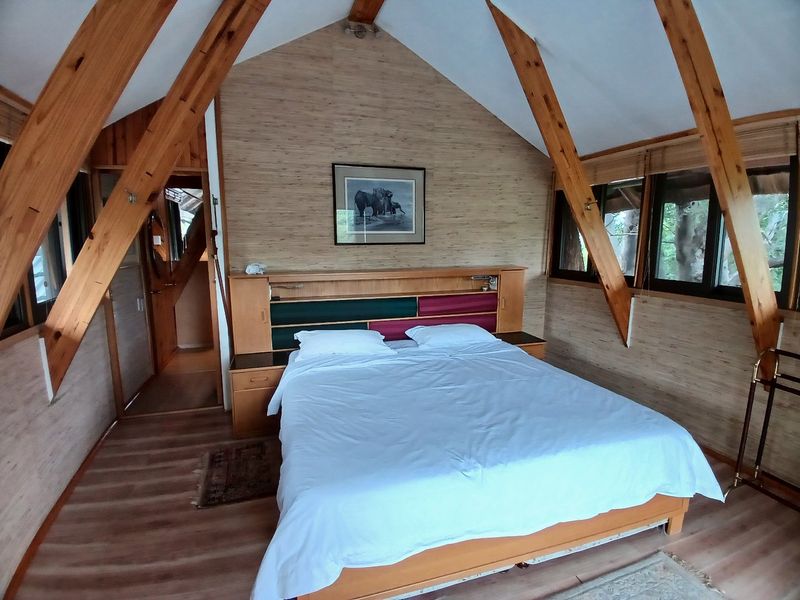 A rustic bedroom featuring a vaulted ceiling with exposed timber rafters, built-in headboard, and laminate flooring with large windows.