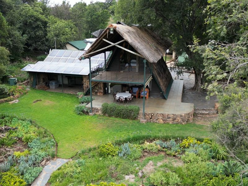 Aerial view of a rustic farm property featuring a unique A-frame thatched structure, solar panel installation, and a spacious stone-paved patio overlooking the garden.