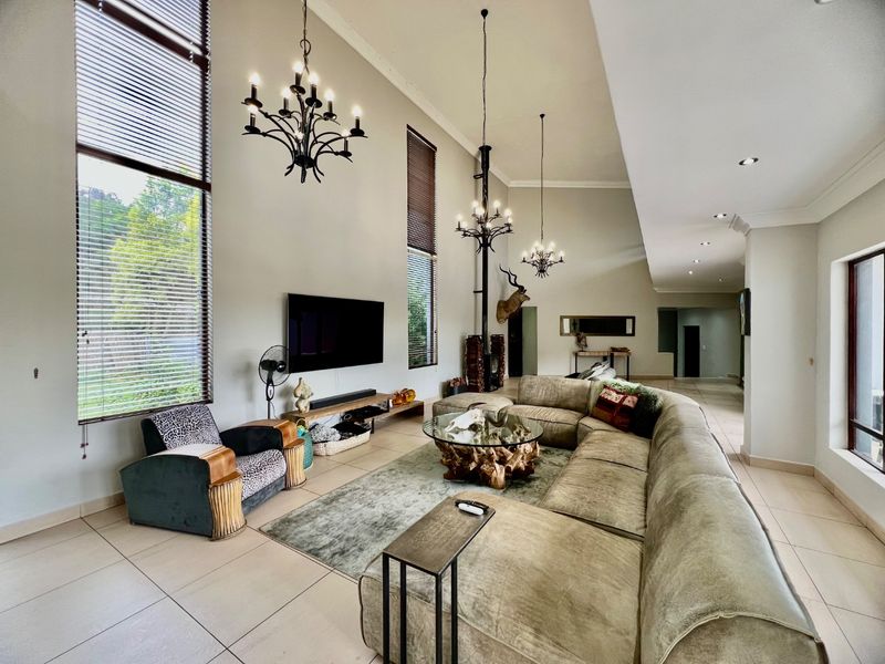 Spacious living room featuring double-volume ceilings, large windows with blinds, elegant wrought iron chandeliers, and a prominent freestanding fireplace.