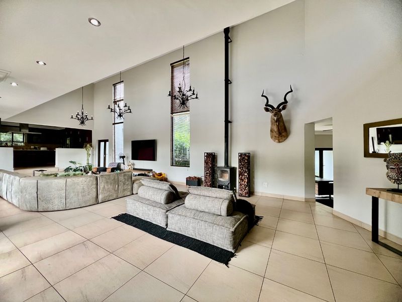 Spacious open-plan living area featuring a vaulted ceiling, tiled flooring, tall windows, and a prominent wood-burning fireplace.