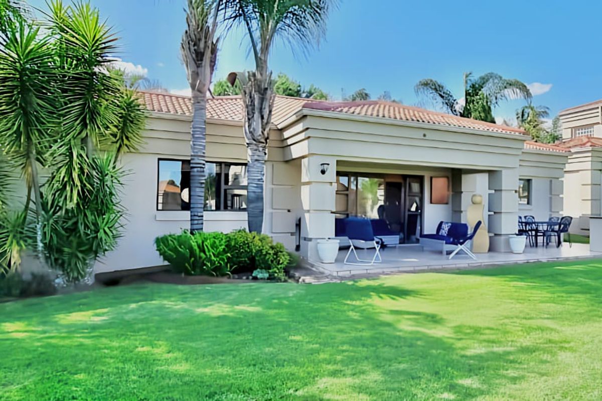 A well-maintained rear exterior view featuring a spacious covered patio, built-in braai area, and a neatly landscaped garden with mature palm trees.