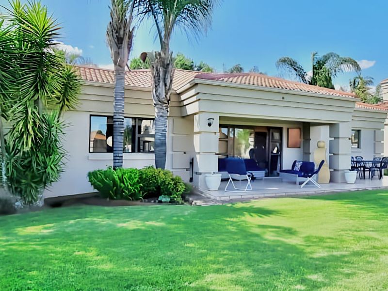 A well-maintained rear exterior view featuring a spacious covered patio, built-in braai area, and a neatly landscaped garden with mature palm trees.