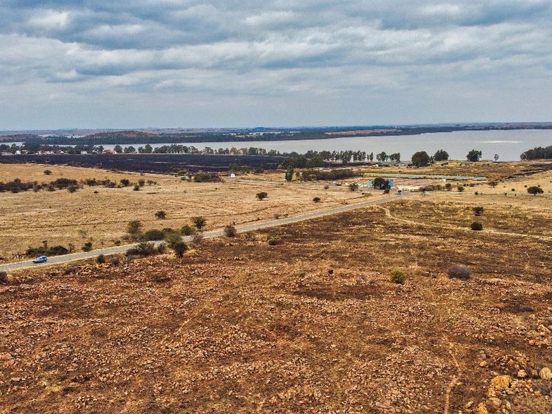 An aerial view of a large, undeveloped residential land parcel featuring natural terrain, gravel access paths, and proximity to a nearby lake.