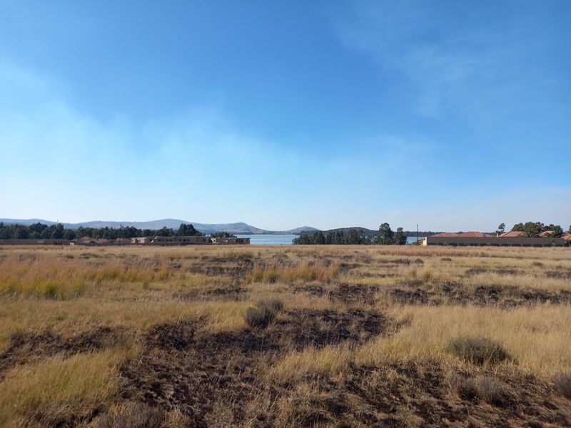 A wide-angle view of vacant residential land featuring natural grass terrain with distant water and mountain views under a clear blue sky.